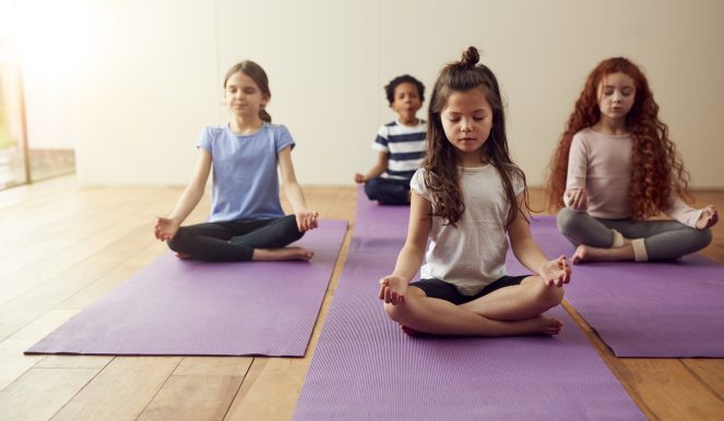 Group Of Children Sitting On Exercise Mats And Meditating In Yoga Studio